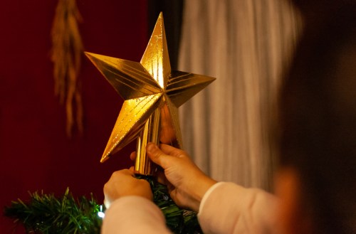 A person places a golden star tree topper on a Christmas tree, with festive decorations in the background.