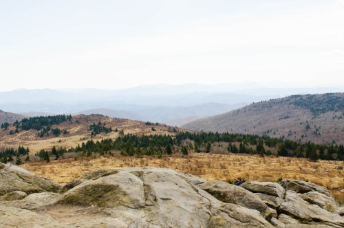 A panoramic view of rolling mountains and valleys, with rocky foreground and sparse trees under a cloudy sky.