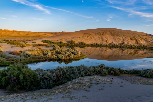 A serene desert landscape with a calm lake reflecting the surrounding dunes and greenery under a clear blue sky.
