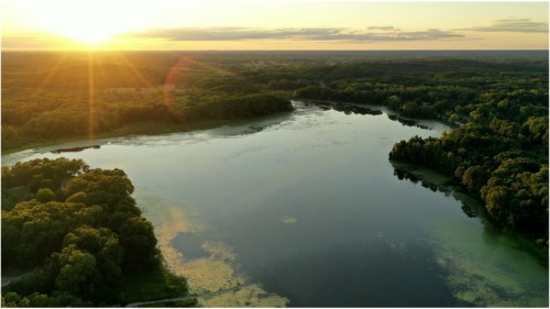 A serene landscape at sunset, featuring a calm lake surrounded by lush greenery and distant hills.