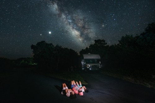 Three people lie on a road, gazing up at a starry sky with the Milky Way, beside a parked camper van.