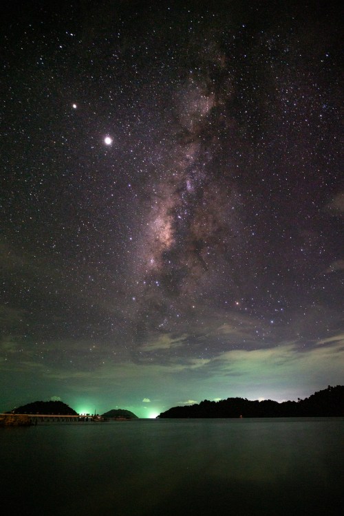 A stunning night sky filled with stars and the Milky Way over a calm sea, with faint green lights on the horizon.