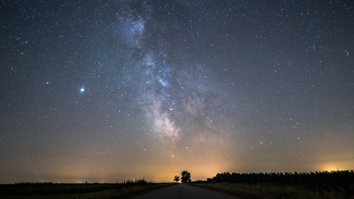 A starry night sky with the Milky Way visible above a dark road lined by trees and distant lights on the horizon.