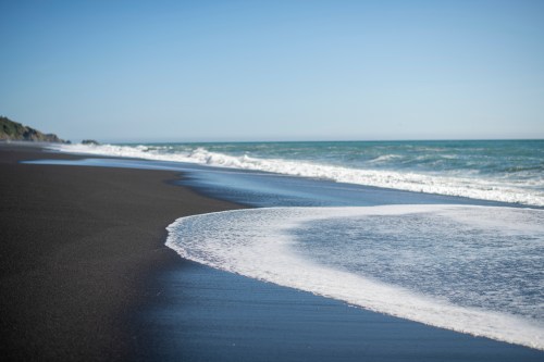 A serene beach scene with black sand, gentle waves, and a clear blue sky.