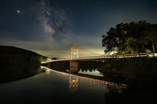 A suspension bridge illuminated at night, reflecting in a calm river under a starry sky with the Milky Way visible.