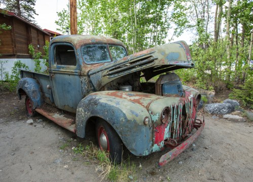 An old, rusty pickup truck with a raised hood, surrounded by greenery and gravel.