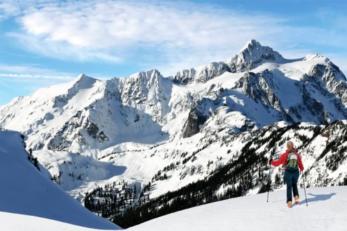 A person hiking on a snowy mountain trail with majestic peaks in the background under a clear blue sky.