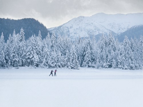 Two people ice skating on a frozen lake, surrounded by snow-covered trees and mountains under a cloudy sky.