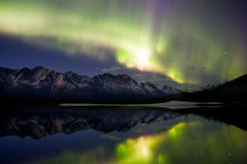 A stunning display of the Northern Lights over snow-capped mountains, reflecting in a calm lake at night.