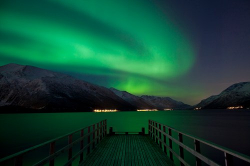 A serene lake scene at night with vibrant green auroras dancing over snow-capped mountains and a wooden pier.