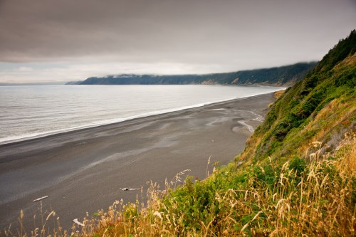 A serene coastal view featuring a black sand beach, cliffs, and overcast skies, with gentle waves lapping at the shore.