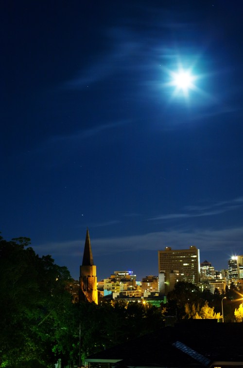 A city skyline at night with a bright moon and a church steeple illuminated against a dark blue sky.
