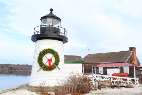 A white lighthouse decorated with a wreath and red bow, beside a calm water body and a rustic building.