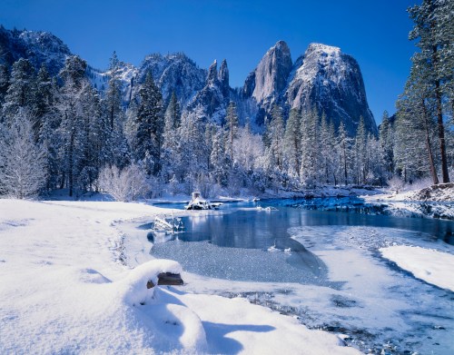 A serene winter landscape featuring snow-covered trees, a frozen river, and majestic mountains under a clear blue sky.