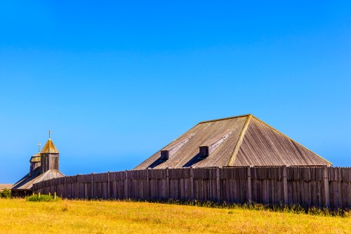 A wooden building with a steeple, surrounded by a tall fence, set against a clear blue sky and grassy field.