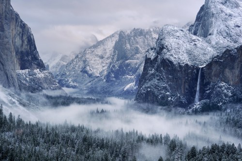 Snow-covered mountains rise above a misty valley, with a waterfall cascading down rocky cliffs in a serene winter landscape.
