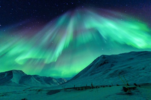 Vibrant green auroras dance across a starry night sky over snow-covered mountains.