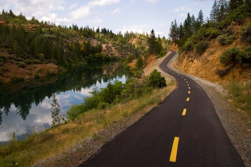 A winding road alongside a calm river, surrounded by lush greenery and trees under a partly cloudy sky.