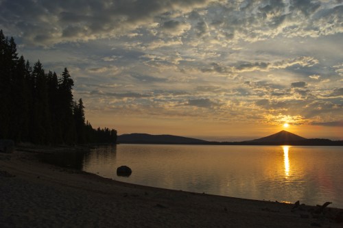 Sunset over a calm lake, with silhouetted trees on the shore and mountains in the background under a cloudy sky.