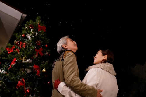 A couple stands together, gazing up at falling snow near a decorated Christmas tree at night.