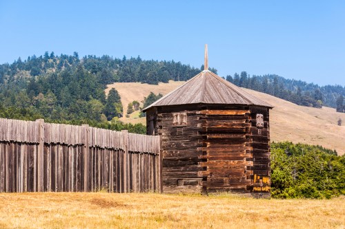 A wooden structure with a pointed roof stands beside a tall wooden fence, set against a backdrop of rolling hills and trees.