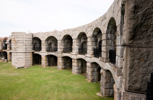 Stone ruins of an ancient structure with arches, set in a grassy area under a cloudy sky.