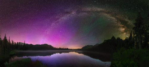 A stunning night sky filled with stars and a colorful aurora, reflecting on a calm lake surrounded by trees.