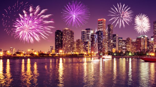 Colorful fireworks illuminate a city skyline at night, reflecting on the water below.