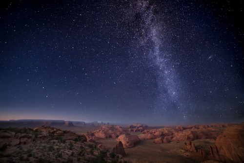 A starry night sky over a desert landscape, with the Milky Way visible and rocky formations in the foreground.