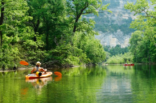 A person in a kayak paddles through a serene, green landscape with cliffs in the background and calm water.