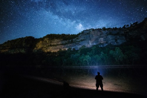 A person stands by a river at night, illuminated by stars and the Milky Way above a rocky cliff and trees.