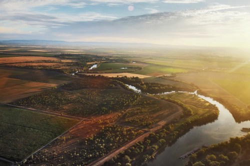 Aerial view of a winding river surrounded by lush fields and trees under a bright sky at sunset.