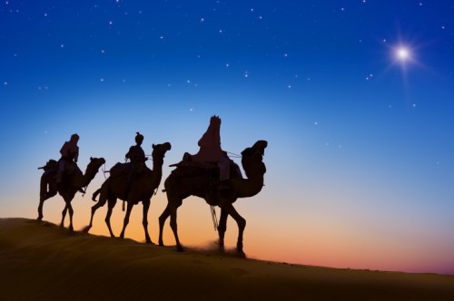 Silhouettes of camels and riders against a starry sky at dusk in a desert landscape.