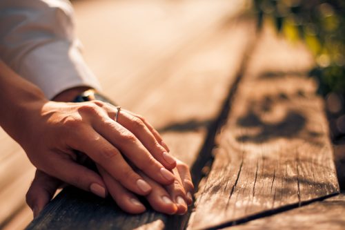 A close-up of two hands gently resting on a wooden surface, one adorned with a ring, in soft, warm lighting.