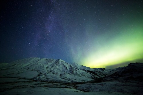 A snowy mountain landscape under a starry sky, illuminated by green auroras in the distance.