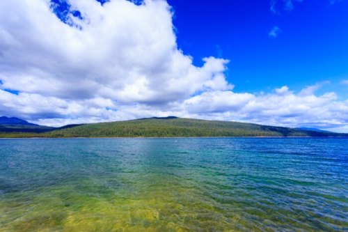 A serene lake with clear blue water, surrounded by lush green hills and a bright, partly cloudy sky.