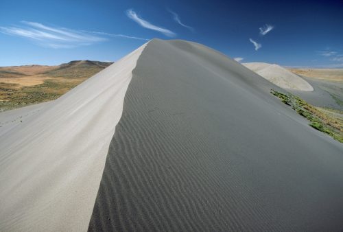 A sweeping view of a sand dune with contrasting light and dark sands under a clear blue sky with wispy clouds.