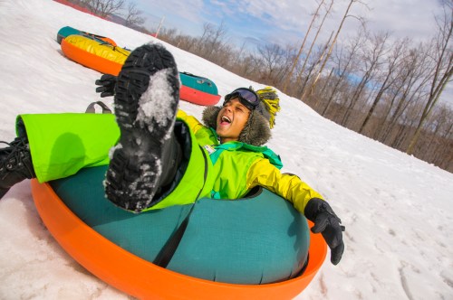 A person in a bright green snowsuit joyfully sliding down a snowy hill on a tube, with trees in the background.