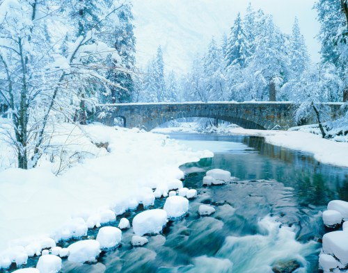 A serene winter scene featuring a snow-covered bridge over a flowing river, surrounded by frosted trees.