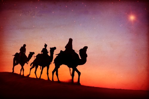 Silhouettes of camels and riders against a colorful sunset in a desert landscape.