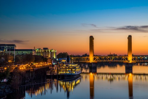 A sunset view of a bridge with illuminated towers, reflecting in the water, alongside a docked boat and city buildings.