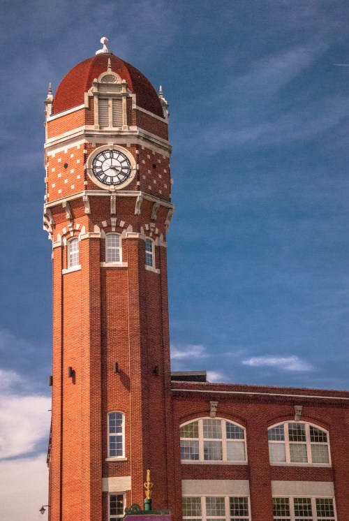 A tall brick clock tower with a red dome against a blue sky, featuring large windows and decorative details.