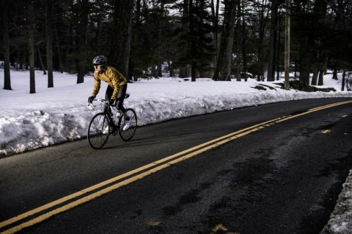 A cyclist in a yellow jacket rides on a snowy road surrounded by trees.