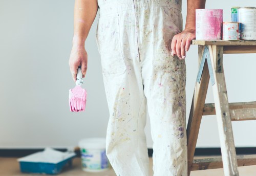 A person in paint-splattered overalls holds a pink paint roller next to a ladder and paint cans in a room.