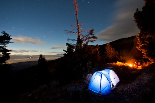 A blue tent illuminated at night, surrounded by trees and a distant campfire under a starry sky.