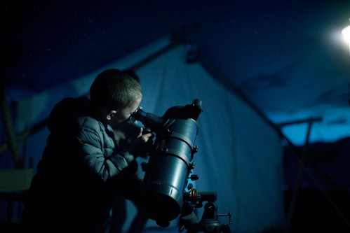 A child looks through a telescope at night, with a tent and starry sky in the background.