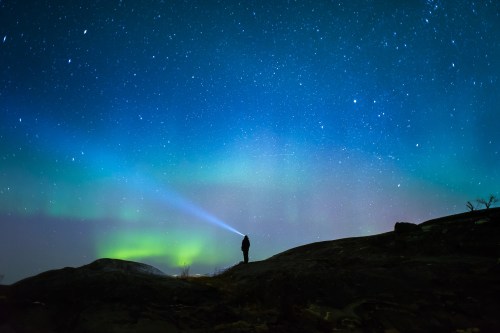 A silhouette of a person with a flashlight stands on a rocky landscape under a starry sky and colorful auroras.