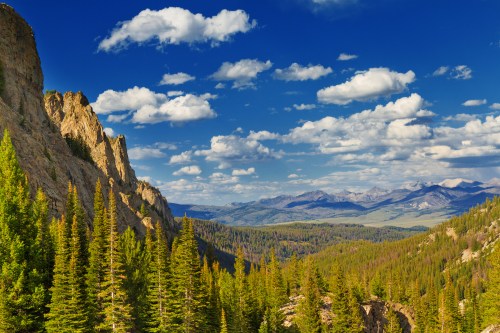 A scenic view of mountains and lush green forests under a blue sky with fluffy white clouds.