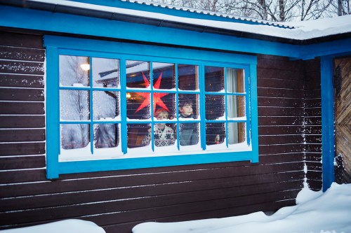 Two children look out a snowy window, framed by blue wood, with a bright red star decoration visible inside.