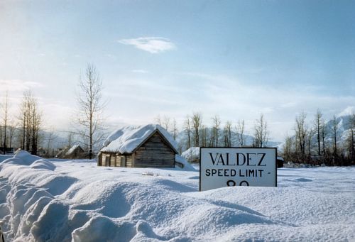 A snowy landscape with a speed limit sign for Valdez and a small, snow-covered cabin in the background.
